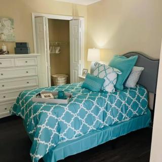 A cozy bedroom with a teal and white patterned bedspread, matching pillows, and a bedside lamp.