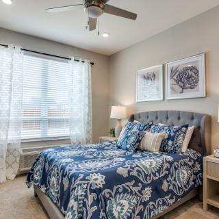 A cozy bedroom featuring a bed with blue patterned bedding, artwork above the headboard, and natural light coming through a window with white curtains.