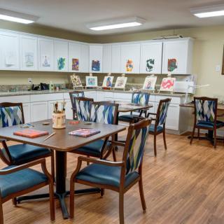 A cozy room with multiple tables and chairs, decorated with artwork and equipped with kitchen cabinets.