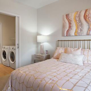 A cozy bedroom with a laundry area visible, featuring a bed, lamp, and artwork on the wall.