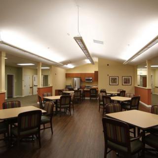 A spacious dining area featuring multiple tables and chairs, well-lit with ceiling lights and adjacent to a kitchen space.