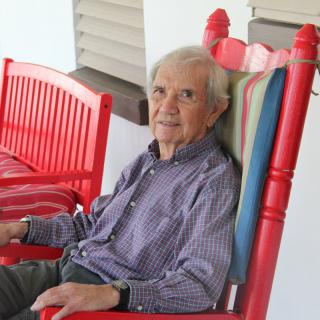 An elderly man sitting comfortably on a red rocking chair outside.