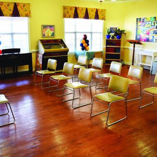 A small classroom setup with chairs arranged for a lecture, featuring a wooden floor and windows letting in natural light.