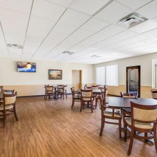 An empty dining room with wooden floors and several tables and chairs arranged neatly, featuring a wall-mounted television.