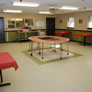 A spacious room featuring multiple tables, a kitchenette area, and bright lighting.