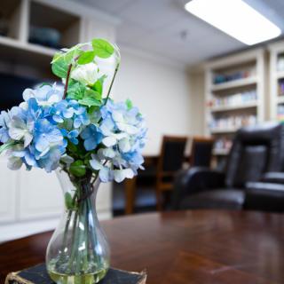 A cozy office setting with a vase of blue and white flowers on a wooden table, surrounded by bookshelves and chairs.