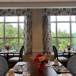 A well-decorated dining room with tables set for a meal, featuring large windows and floral centerpieces.