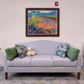 A cozy living space featuring a light gray sofa with colorful cushions and a teddy bear, complemented by a vibrant painting on the wall.