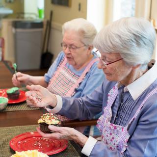 Two elderly women enjoying decorating cupcakes in the kitchen.