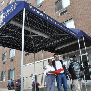 A group of people standing in front of a building with an awning labeled 'Madison York' holding a book.