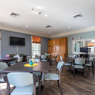 A well-lit community room with several tables and chairs, a television, and decorative elements.