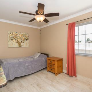 A cozy bedroom with a single bed, wooden nightstand, and a ceiling fan, featuring beige walls and red curtains.