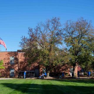 A brick building with an American flag in front, surrounded by trees and a green lawn under a clear blue sky.