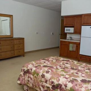 A cozy bedroom with a floral bedspread, wooden dresser with mirror, and a kitchenette featuring a refrigerator and microwave.