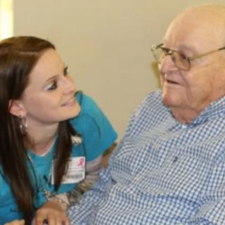 A caregiver interacts warmly with an elderly man, both smiling.