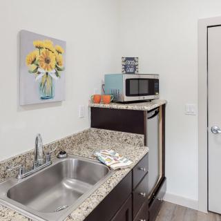 A small kitchen area with a granite countertop, featuring a sink, microwave, and a painting of flowers on the wall.
