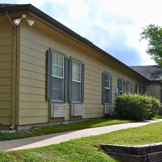 A single-story building with yellow siding, multiple windows, and surrounding greenery.