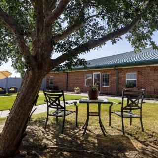 A tranquil backyard setting featuring a small patio table with chairs under the shade of a tree, adjacent to a brick building.