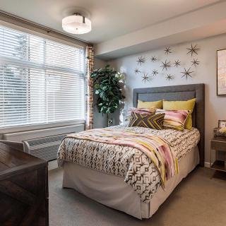 A modern and cozy bedroom with stylish decor, featuring a bed with patterned bedding, a large window allowing natural light in, and decorative elements on the wall.