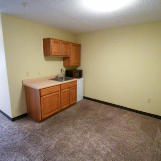 A small kitchenette with wooden cabinets, a sink, and carpeted flooring in a room with yellow walls.