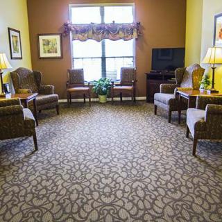 A cozy living room with patterned carpet and several chairs arranged around a window, complemented by warm lighting.
