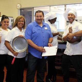A group of chefs and a person holding a certificate, posing in a kitchen with cooking utensils.