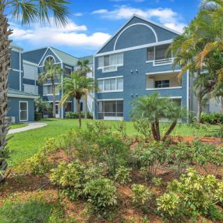 A scenic view of modern apartment buildings surrounded by lush greenery and palm trees under a bright, sunny sky.