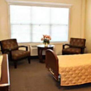 A cozy bedroom featuring two chairs, a bed with a yellow quilt, and a window with blinds.