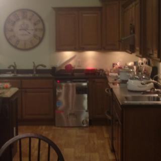 A cozy kitchen with wooden cabinets, a large clock on the wall, and various appliances on the countertop.