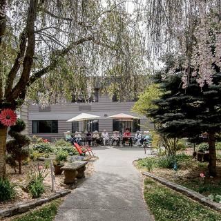 A scenic garden with a curving pathway, surrounded by trees and flowers, leading to an outdoor seating area with umbrellas.
