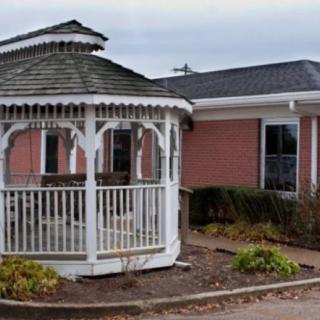 A white wooden gazebo in front of a brick building surrounded by sparse landscaping.