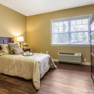 A cozy bedroom with a neatly made bed, a lamp on the nightstand, and a window providing natural light.