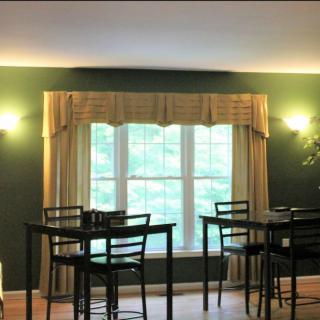 A cozy dining area with dark green walls, featuring multiple tables and chairs, illuminated by wall lamps and natural light from a large window with beige curtains.
