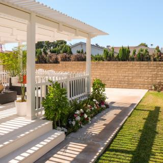 A serene backyard patio with a white pergola, surrounded by lush greenery and a well-manicured lawn.
