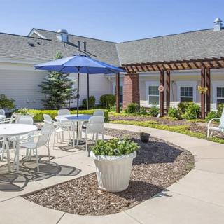 A sunny patio area with tables, chairs, and an umbrella surrounded by a garden and walkway.
