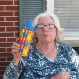 An elderly woman wearing glasses is seated outdoors, holding a colorful water bottle.