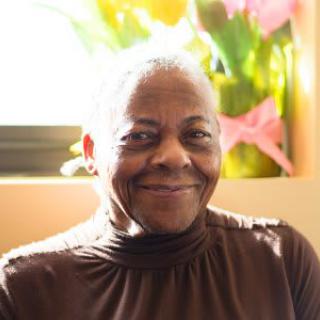 An elderly person smiling warmly indoors with soft natural light coming through the window.