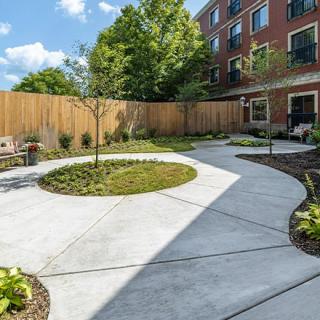 A beautifully landscaped garden with a circular paved path, surrounded by greenery and bordered by a wooden fence, adjacent to a building.