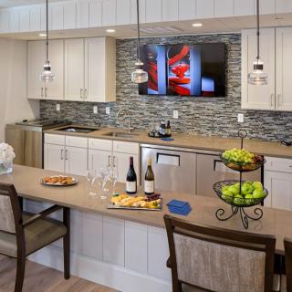 A modern kitchen with a central island, featuring wine bottles, fruit bowl, and a TV above the tile backsplash.