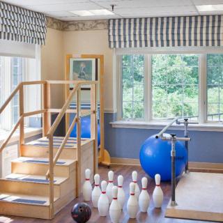 A physical therapy room with exercise equipment including stairs, a stability ball, parallel bars, and bowling pins near large windows.