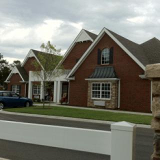 A suburban brick house with a neatly maintained lawn and a white fence under a cloudy sky.