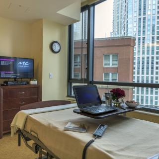 A hospital room featuring a patient bed with a laptop and flowers, overlooking an urban cityscape through large windows.