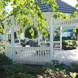 A charming white gazebo surrounded by lush greenery in a garden setting under dappled sunlight.