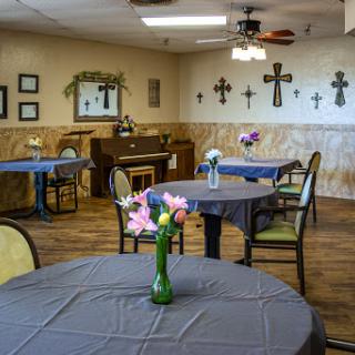 A cozy dining area featuring tables with floral arrangements, a piano, and decorative crosses on the walls.
