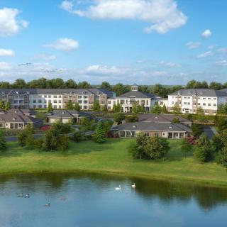 A serene residential community with buildings surrounded by greenery and a calm lake under a clear blue sky.