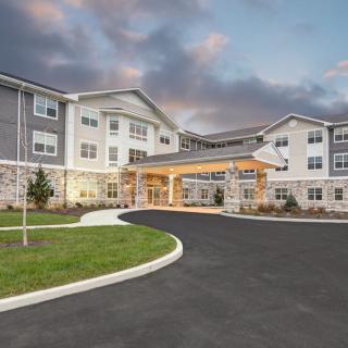 A modern, multi-story building with a driveway and landscaped surroundings under a cloudy sky.