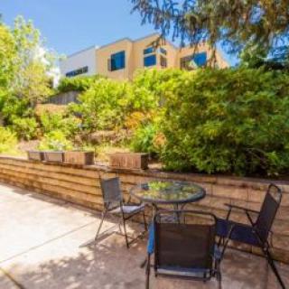 A sunny patio area with a glass table and four chairs, surrounded by lush greenery.