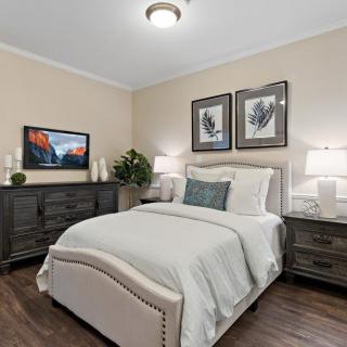 A cozy and elegantly decorated bedroom featuring a plush bed, matching nightstands with lamps, a dresser with a television, and framed artwork on the walls.