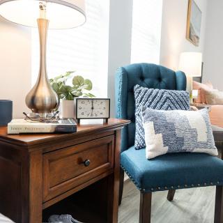 A cozy living room corner featuring a blue upholstered chair with decorative pillows, next to a wooden side table adorned with a lamp and small clock.