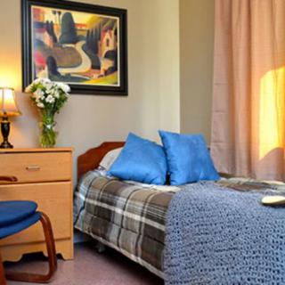 A cozy bedroom with a neatly made bed, blue pillows, and a side table featuring a lamp and flowers.